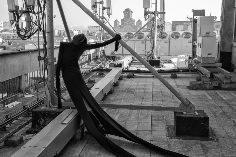 TESKOBA: figure in a fitted black bodysuit with long trailing panels, leaning on a rooftop steel beam