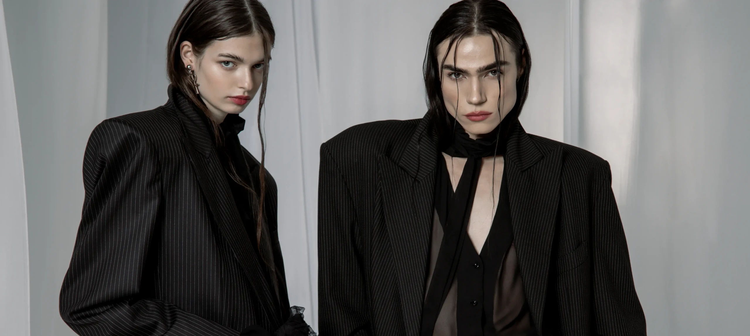 Two models in black oversized pinstripe blazers, one with a sheer shirt and a neck scarf