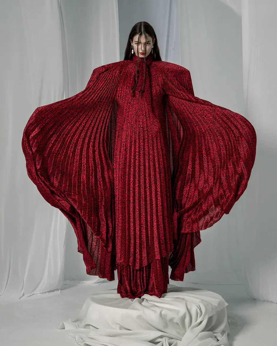 Model in a red printed pleated gown with sweeping wing-like sleeves, standing on draped fabric