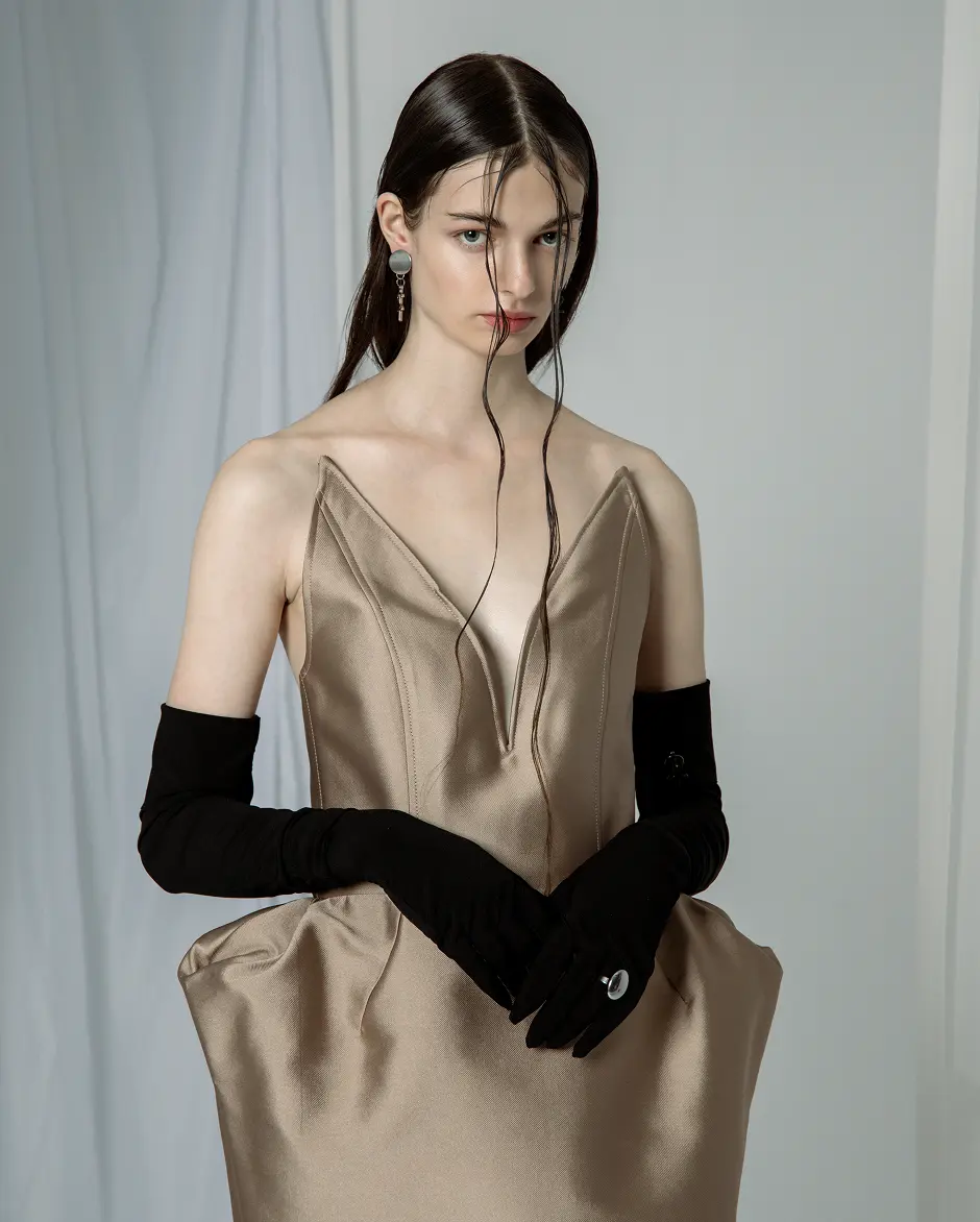 Model in a strapless beige satin dress with a deep V neckline, sculpted hips and long black gloves