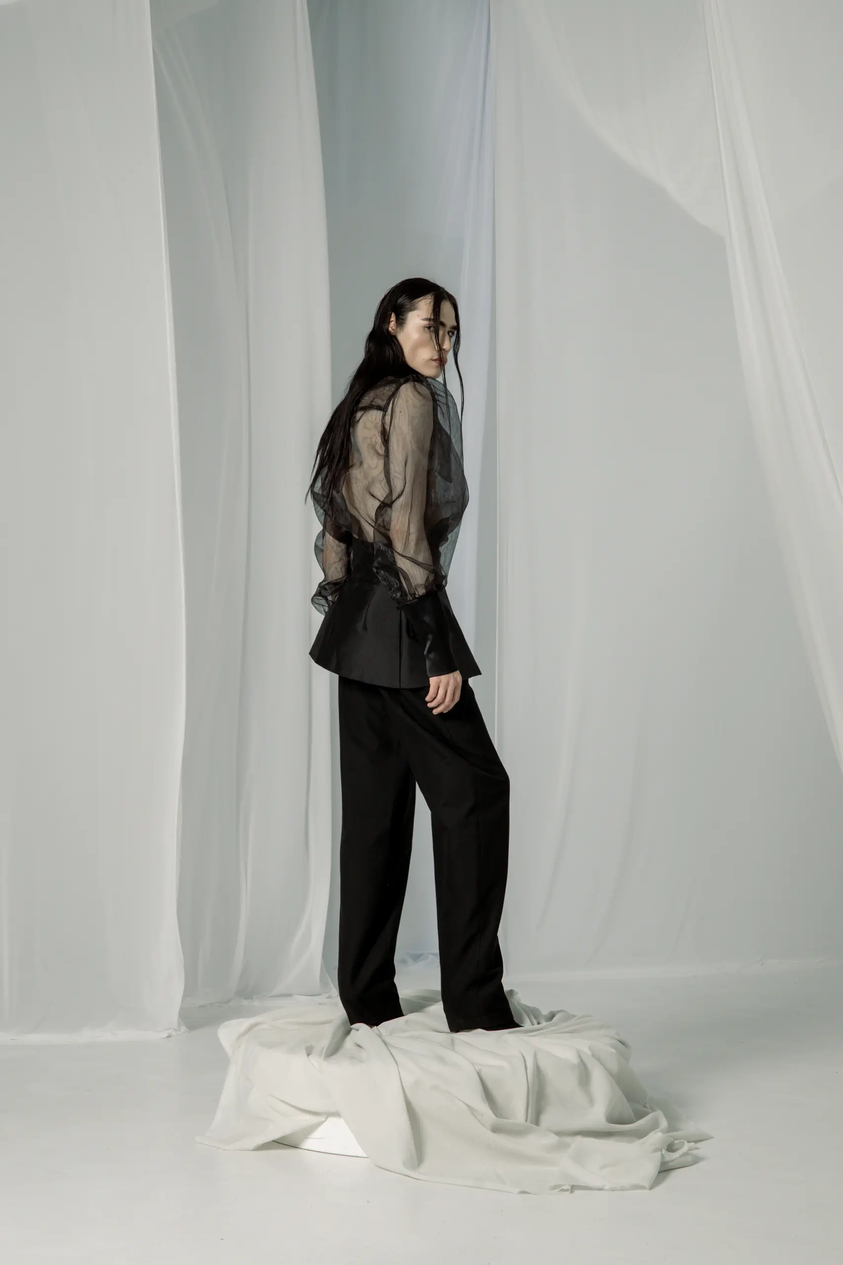 Model gleda preko ramena, providna bluza od organze i crne pantalone, pogled s leđa
