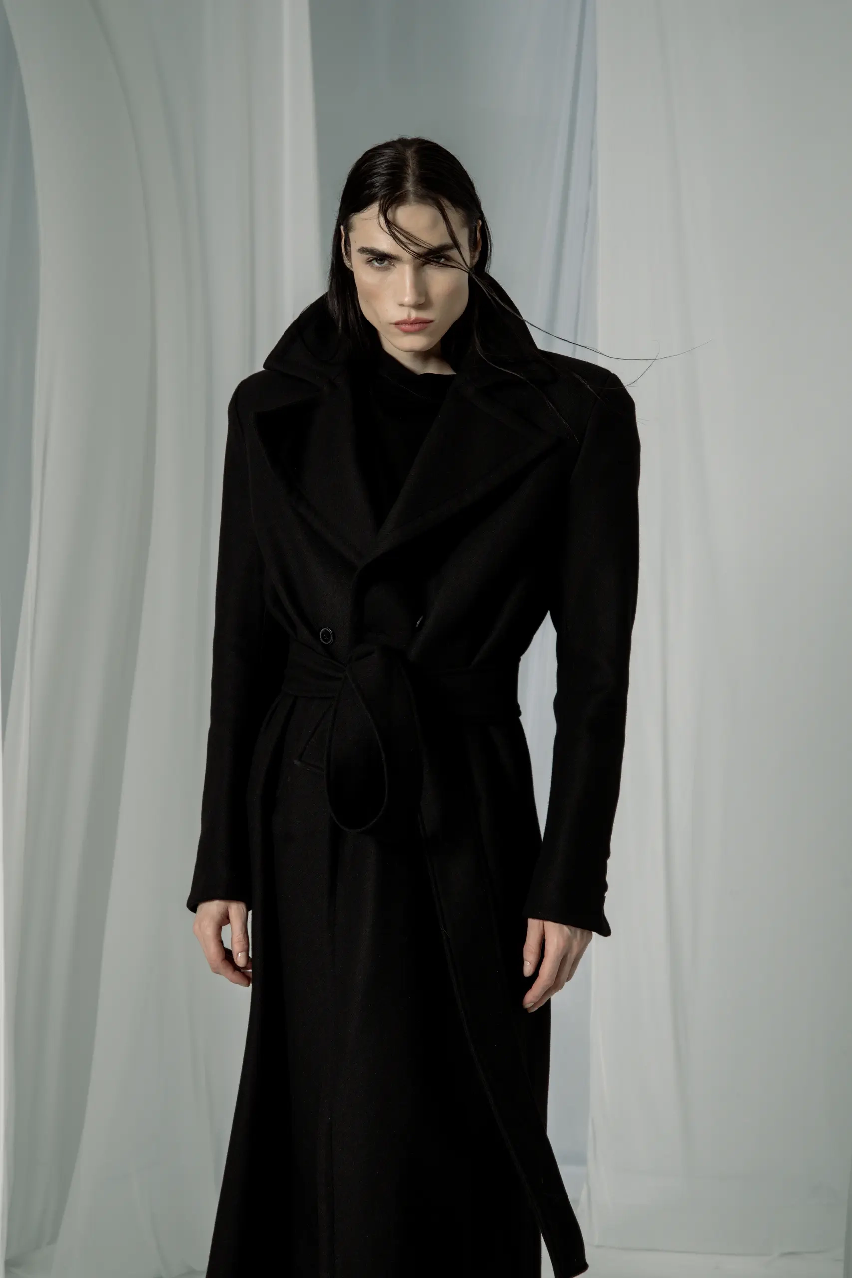 Black double-breasted coat with wide lapels and a tie belt, waist-up shot with windswept hair