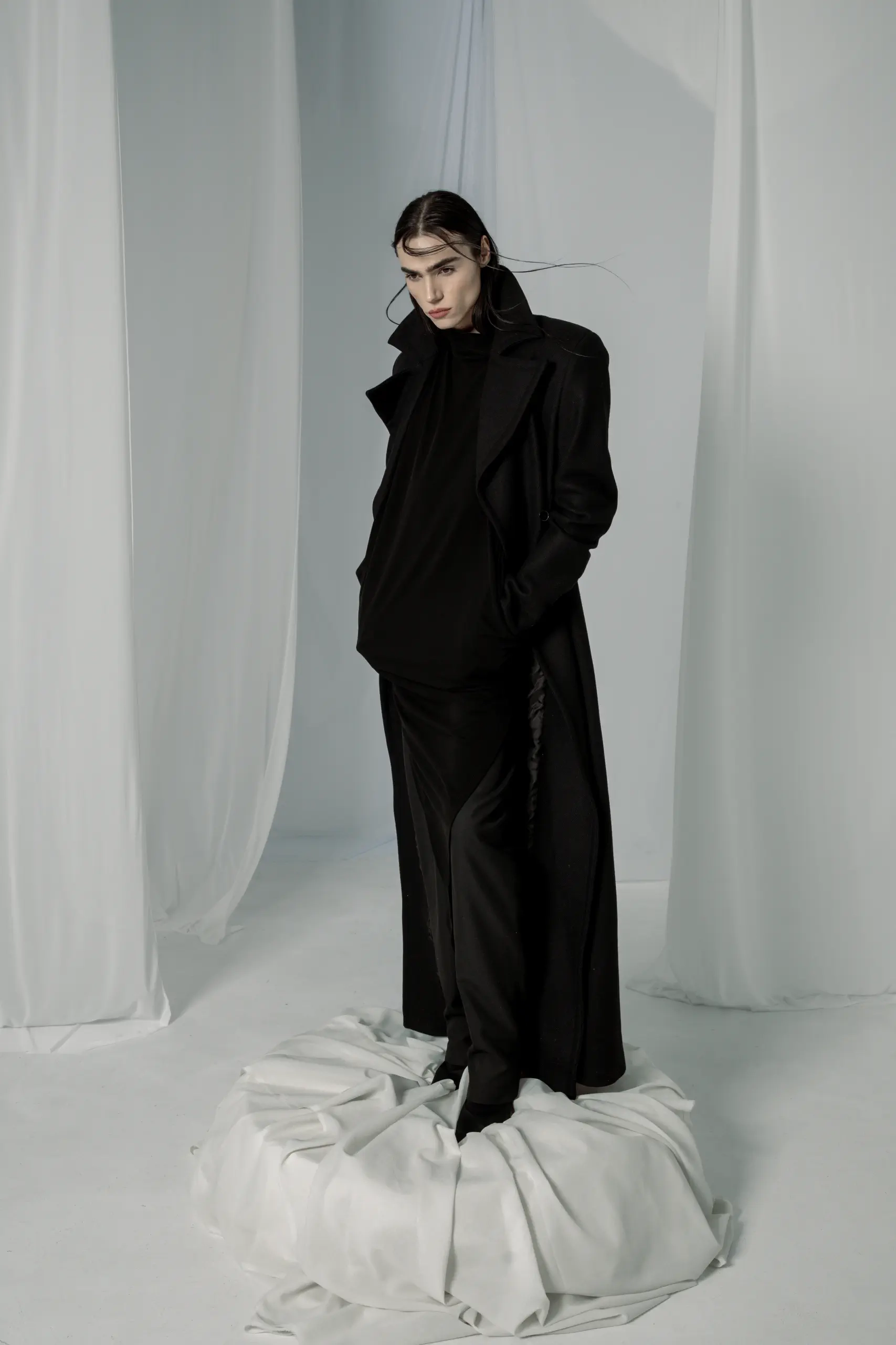 Model u dugom crnom oversize kaputu preko satenskih širokih pantalona, ruke u džepovima, na draperiji