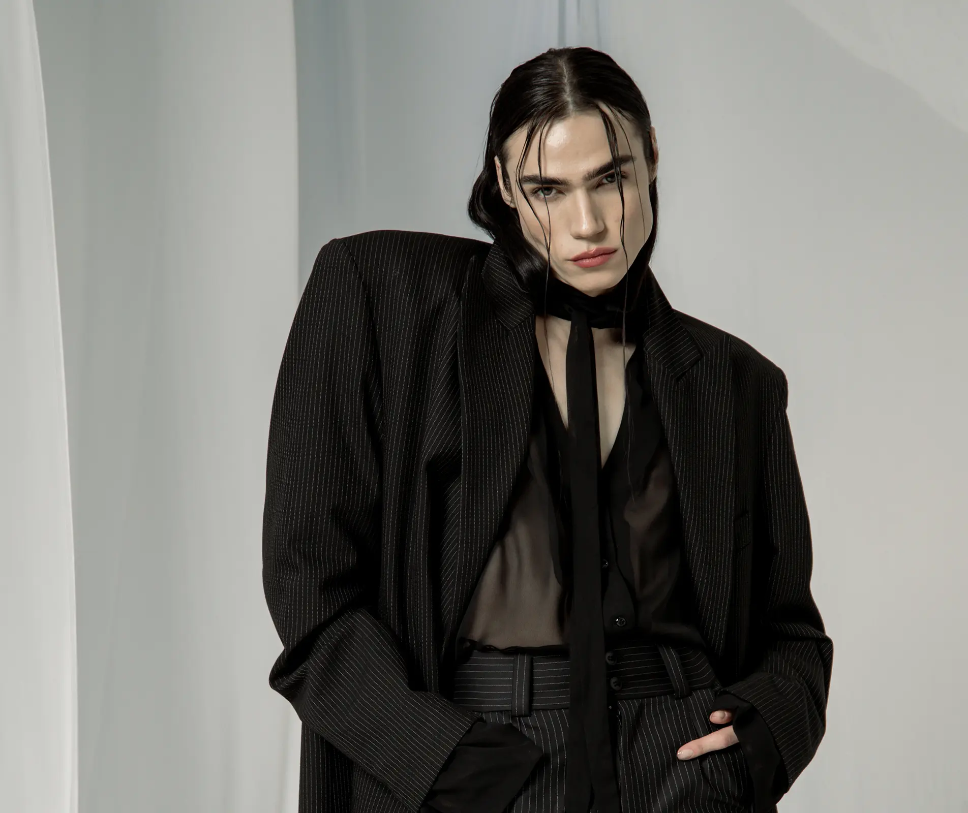 Model in an oversized pinstripe suit with a sheer black shirt and long neck scarf, hand in pocket